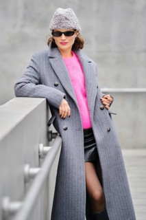 A woman stands confidently on an urban street, dressed in a long gray coat over a vibrant pink
