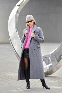 A woman in a long gray coat and pink sweater posing near a large modern sculpture. She wears black