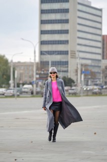 A stylish woman walks along a city sidewalk, wearing a vibrant pink sweater, leather skirt and a