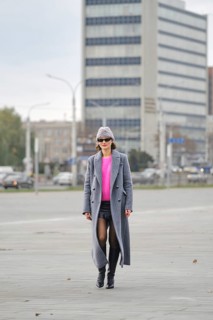 A stylish woman walks through an urban setting wearing a gray long coat and a bright pink sweater