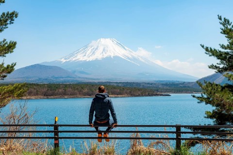 Young man sitting on railings next to a road and looking across the lake to Mt Fuji volcano, Motosu