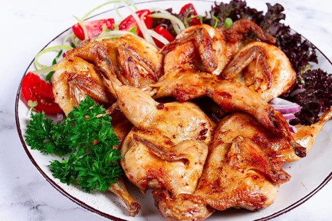 Baked quail, served on a plate with crispy lettuce leaves, fresh tomatoes and herbs, on a white
