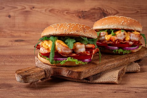 Two shrimp burgers and buns with sesame seeds, bacon, lettuce, tomatoes and onions, on a wooden