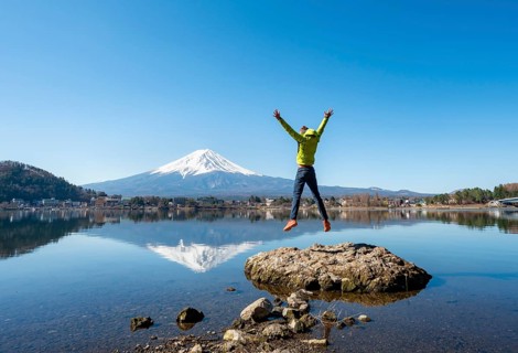 Young man standing on a rock in water, jumping into the air, volcano Mt. Fuji is reflected in Lake