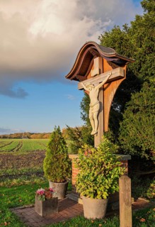 Wooden cross with Jesus figure against the backdrop of a rural landscape in sunlight, Münsterland,