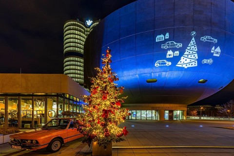 Illuminated Christmas tree with red balls, next to a historic BMW vehicle, in front of the BMW