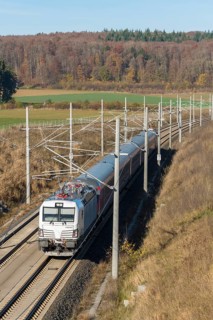 A passenger train travels through an autumnal landscape with fields and forests in the background,