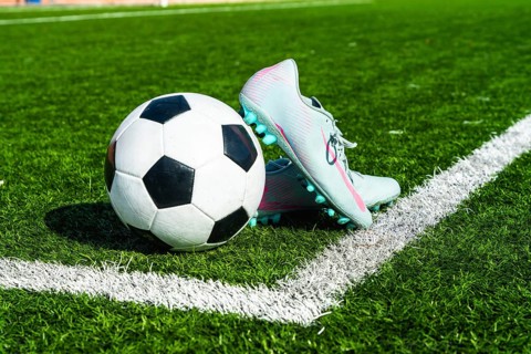 Soccer ball and football cleats are sitting on a green artificial grass pitch, close to the white