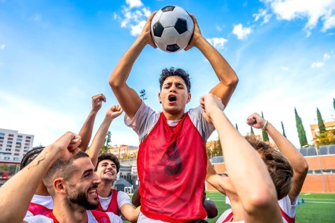 Young men soccer teammates in red bibs celebrate victory, raising a ball overhead with joyful