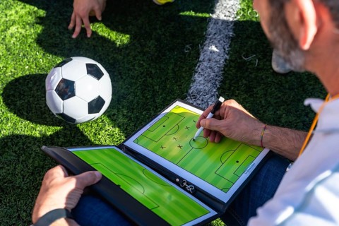 Soccer coach sketching tactical game plan on a clipboard beside a ball on artificial grass near a