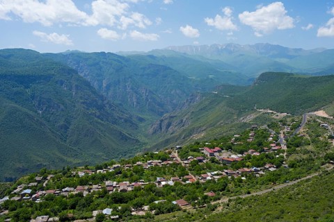 A small village is situated in a green valley surrounded by forested mountains under a cloudy sky,