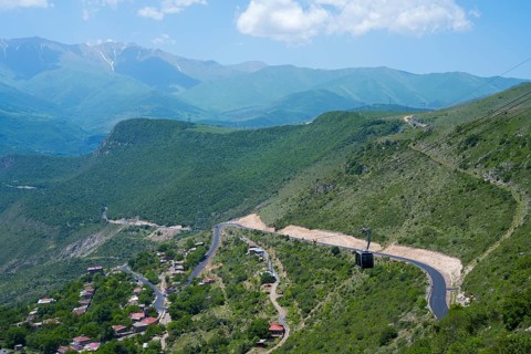 A winding road runs through green hills and passes a small village, with views of mountains, Tatev