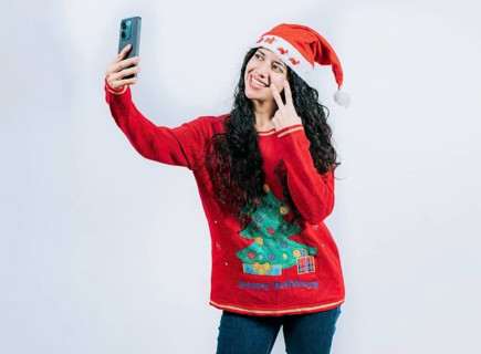 Funny girl in Christmas hat and sweater taking selfie, isolated. Happy young latin woman in