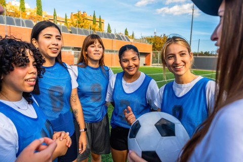 Diverse women football players smiling and listening, holding a soccer ball on a green artificial