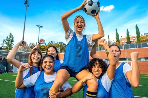Group of diverse women soccer players cheering and lifting a teammate with a soccer ball on a green