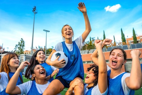 Joyful female soccer players lifting a teammate with the ball overhead, celebrating a win together