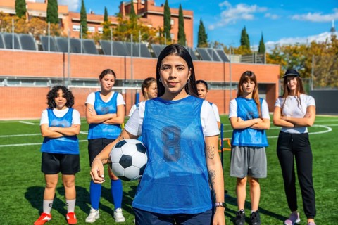 Young female soccer players standing on a sunny artificial turf field, with one player holding a