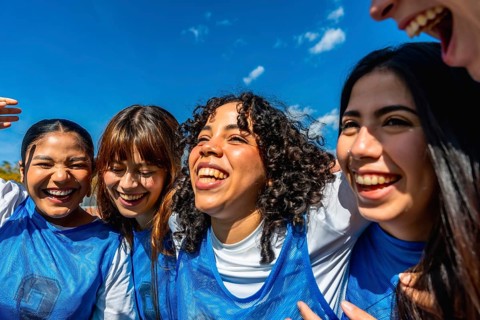 Diverse group of young girls standing close, smiling and laughing under a bright blue sky,