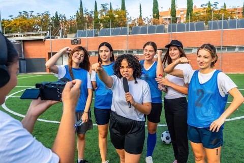 Reporter with microphone interviewing a diverse women's soccer team on a green field, capturing