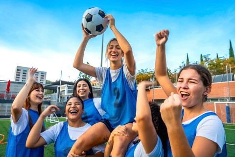 Young happy women soccer team players celebrating victory, one player is sitting on teammates'