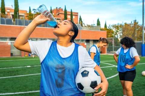 Young woman soccer player in a blue bib holding a football, refreshing herself by drinking a sports