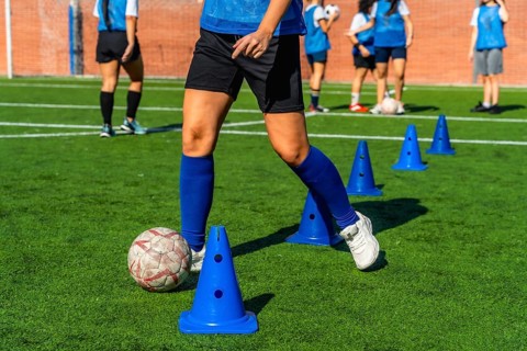 Female football player dribbling a soccer ball between blue training cones on a green artificial