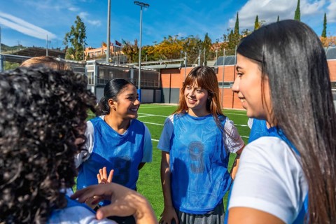 Female soccer teammates in blue bibs smiling and chatting on a sunny field, sharing a light moment