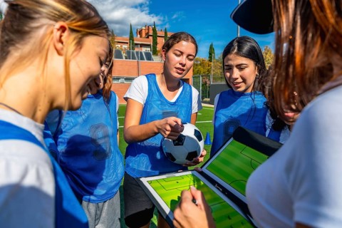 Women's soccer coach and team players discussing game strategy on a clipboard with a soccer ball,