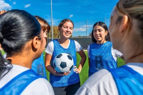 Women soccer players in blue uniforms huddled on a green field, smiling and planning strategy