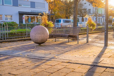 Public bench and large sphere on paved square, autumn trees and buildings in the background,