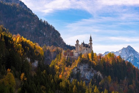 Neuschwanstein Castle rises on a hill covered with autumn trees, mountains in the background,