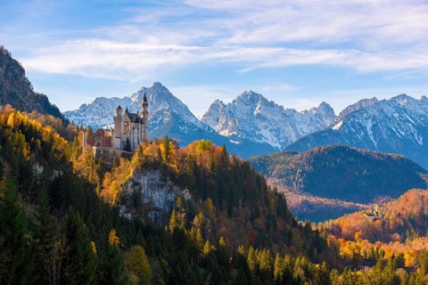 Neuschwanstein Castle in a forest of colorful autumn trees, against snow-covered mountains and blue