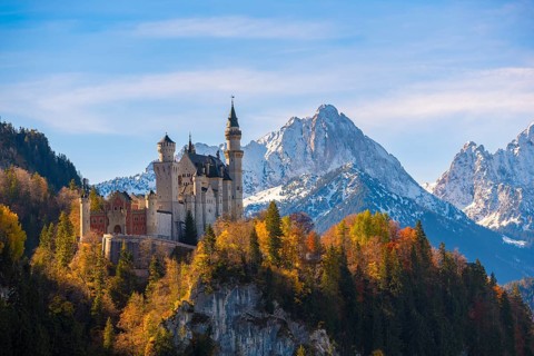 Neuschwanstein Castle rises impressively against a snowy mountain backdrop, surrounded by autumnal