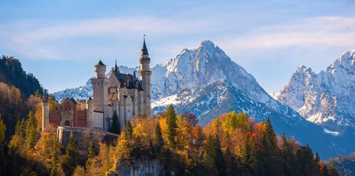 Neuschwanstein Castle is perched on a hill in front of snow-covered mountains, surrounded by