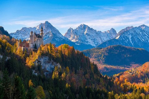 Neuschwanstein Castle stands against a picturesque backdrop of snow-capped mountains and autumn