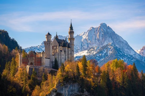 The picturesque Neuschwanstein Castle between autumnal forest and dramatic snowy mountain ranges of