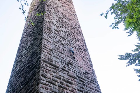An athlete climbs up a high stone wall surrounded by trees under a clear sky, Burgruine Mandelberg,