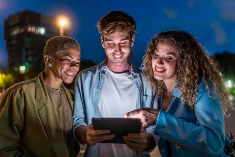 Three smiling diverse friends standing outdoors at night, looking at a shared digital tablet screen