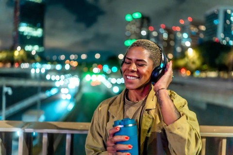 Woman standing on a bridge, holding a travel mug and wearing headphones while happily listening to
