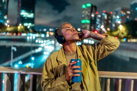 Woman standing on an overpass bridge in a city at night. Wearing headphones and holding a beverage