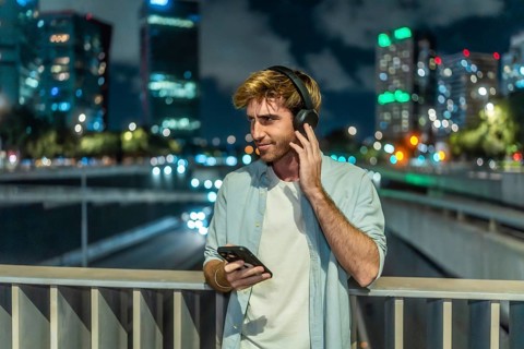 Young man is standing on a bridge, listening to music through headphones and holding a smartphone,