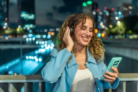 Woman with headphones and smartphone enjoying music while smiling cheerfully, standing on a bridge