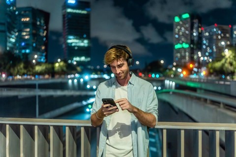 Young man wearing headphones and casual clothing standing on a city bridge at night, smiling and
