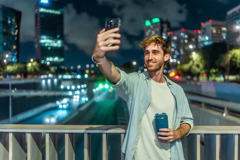 Young man smiling and taking a selfie with a smartphone, holding a reusable coffee cup on an