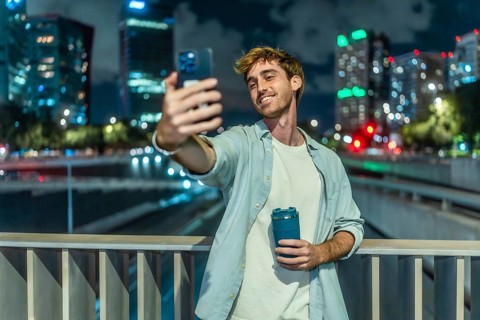 Young man smiling and taking a selfie with a smartphone while holding a reusable coffee cup,