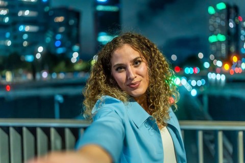 Young woman with curly hair extending her arm to take a selfie, smiling to the camera with blurred