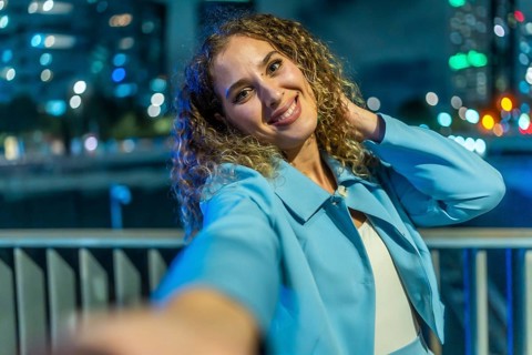 Young woman with curly hair smiling brightly and playfully taking a selfie at night, with a blue