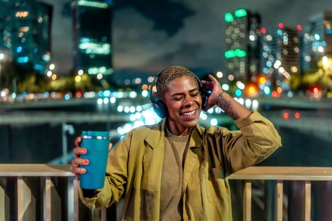 Happy young black woman standing on an urban bridge at night, enjoying music through headphones and