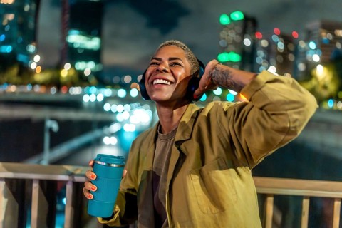 Smiling woman listening to music with headphones, adjusting an earcup, and holding a reusable