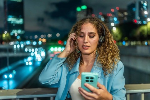 Young woman with curly hair wearing headphones and looking at her smartphone while enjoying music